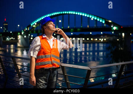 Architect talking on his mobile phone during blue hour by the river with illuminated bridge and city lights in the background Stock Photo