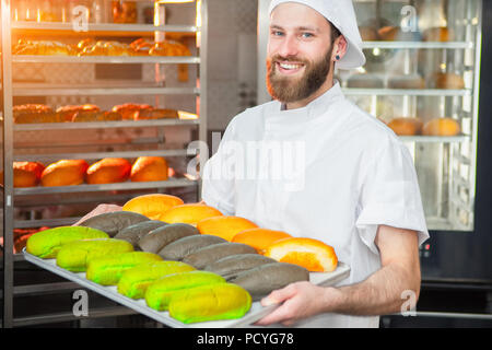 A young handsome baker is holding colorful fresh baked goods on a sheet on the background of the oven. Stock Photo