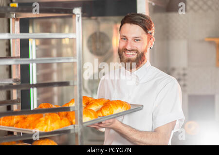 A young handsome baker takes out hot fresh baking from the oven in the bakery. Stock Photo