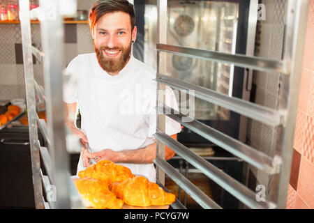 A young handsome baker takes fresh hot croissants from the oven on a shovel. Stock Photo