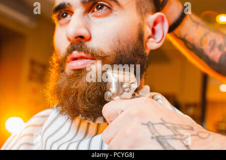 Barber cuts a beard of vintage hair clippers to a young handsome guy with a beard and mustache. Men's hair salon. Stock Photo