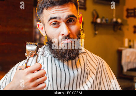 A young handsome guy with a beard and mustache is holding vintage hair clips in his right hand. Men's hair salon. Stock Photo
