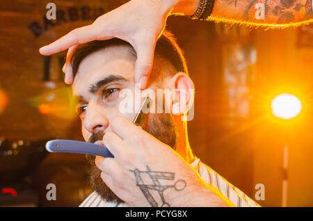 Barber shaves the dangerous razor of a handsome young man with a mustache and beard. Shaving a dangerous razor in a male hairdresser Stock Photo