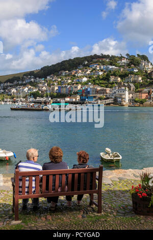 Holiday makers sit on a bench at Bayard's Cove, Dartmouth, looking across the River Dart to Kingswear on a bright summers day Stock Photo