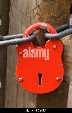 Large bright vibrant red metal padlock holding together a strong silver steel chain links round an old wooden gatepost. Stock Photo
