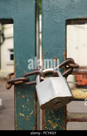 Large old silver steel metal padlock holding together a strong rusting brown chain links round an old green gate with industrial building backgroun Stock Photo