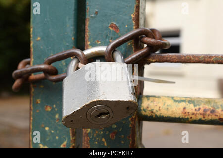 Large old silver steel metal padlock holding together a strong rusting brown chain links round an old green gate with industrial building backgroun Stock Photo