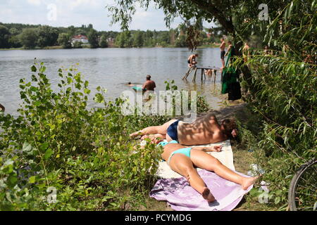 Young Ukrainian woman and man rest on bank of lake in Kiev enjoying delightful summer weather sunbath while other folks on beach fish and have fun Stock Photo