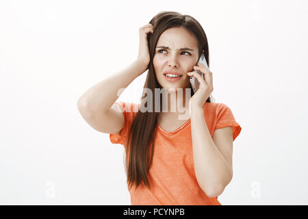 Indoor shot of beautiful urban female student in orange clothes, scratching head and frowning while recalling something, talking on phone, being unsur Stock Photo