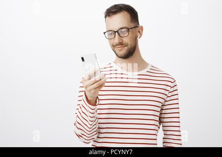 Indoor shot of good-looking relaxed european man in glasses, holding smartphone, picking song to listen, wearing wireless earphone, being pleased with Stock Photo