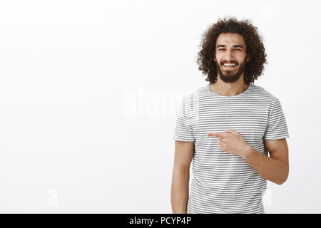 Studio shot of happy carefree handsome man with beard in stylish striped t-shirt, laughing from joy and pointing left with index finger, enjoying spen Stock Photo