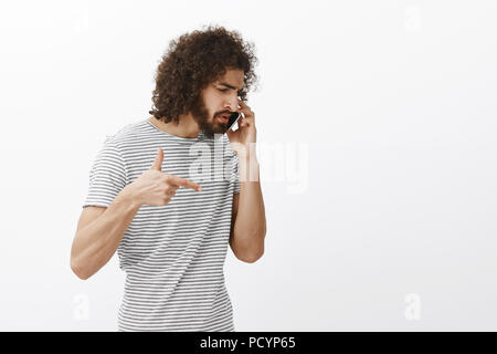 Studio shot of bossy handsome hispanic businessman with beard and afro hairstyle, turning right while talking on smartphone and gesturing with finger  Stock Photo