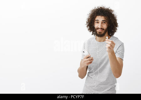 Wanna share earbuds. Studio shot of friendly good-looking slim male model in striped t-shirt, pulling earphone towards camera and holding smartphone,  Stock Photo