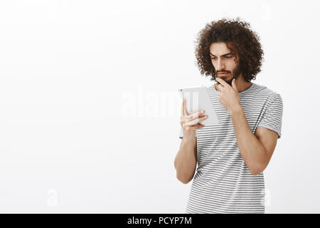 Studio shot of focused busy attractive male with curly hair, holding smartphone, looking at screen thoughtfully and rubbing beard, reading interesting Stock Photo