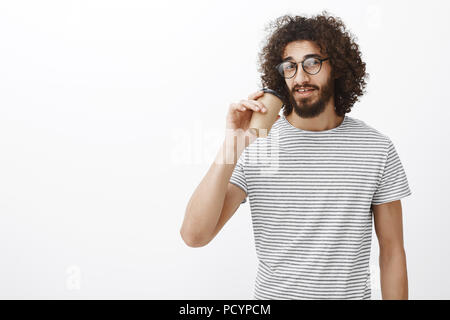 Portrait of confident good-looking slim male barista in trendy t-shirt and eyewear, drinking coffee from cup and gazing at camera, having break and en Stock Photo