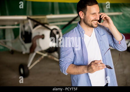 Young handsome young pilot checking airplane in the hangar and using mobile phone Stock Photo