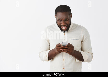 Disappointed upset dark-skinned male model in white shirt, frowning and complaining while reading disappointing message in smartphone, holding device  Stock Photo