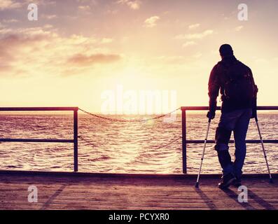 Hurt man with hooded jacket and forearm crutches  standing on sea bridge within  early morning and thinking. Gloomy nostalgic silhouette of sad lonely Stock Photo