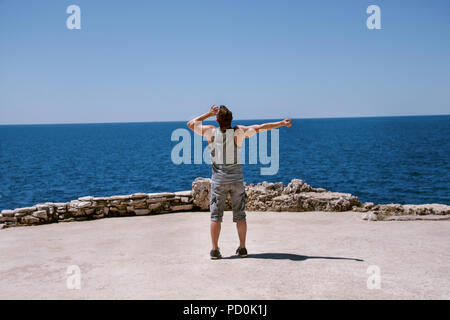 He looks at the sky and shouts to the sea. Tourist man on the rocky shore of the sea. He opened his arms with happiness. Stock Photo