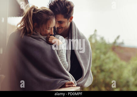 Romantic couple on a winter holiday. Man and woman standing together in a hotel room balcony wrapped in blanket. Couple embracing and smiling.
