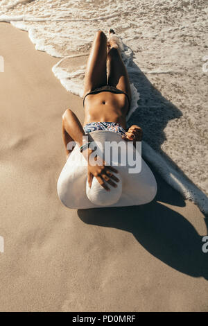 Woman on vacation wearing sun hat relaxing at the beach. Woman in bikini sunbathing lying near the sea shore wearing sun hat with sea waves. Stock Photo