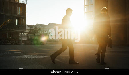 Men commuting to office early in the morning with rising sun in the background. Businessmen with office bag in hand walking on the street to office. Stock Photo