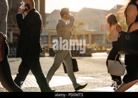 Men and women using mobile phone while commuting to office on a crowded street with sun flare in the background. People leading a busy life using mobi Stock Photo