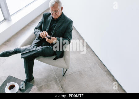 To view of old businessman using with mobile phone in office lobby. Mature corporate professional reading text message on his smartphone during break. Stock Photo