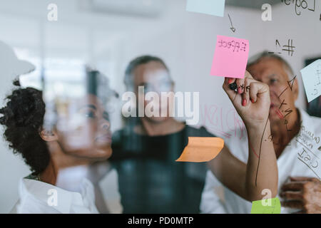Business colleagues working on project together. Business woman writing on sticky note on glass wall with coworkers standing by in office. Stock Photo