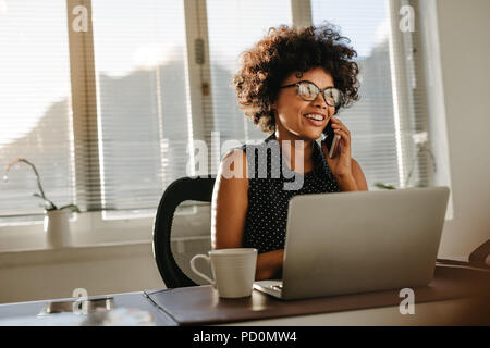 Young african woman working at her desk with laptop and talking on phone. Female wearing casual attire working at a startup office. Stock Photo