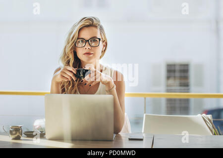 Portrait of attractive young woman sitting at cafe table with laptop and having coffee. Young caucasian businesswoman having coffee at coffeeshop. Stock Photo