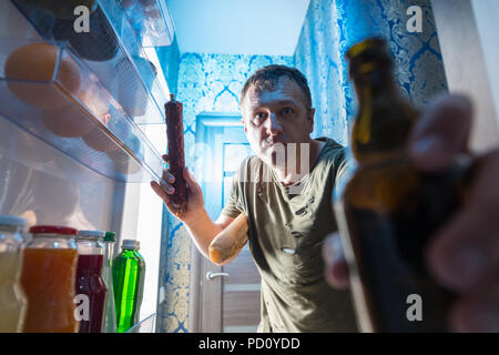 Man grabbing a salami and beer from the fridge viewed from inside the refrigerator looking out in an unusual point of view Stock Photo