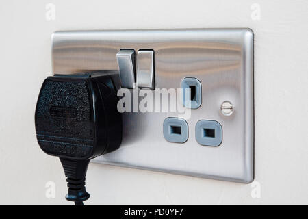 A British three pin plug plugged in to a domestic stainless steel double wall socket switched on. England UK Great Britain Stock Photo