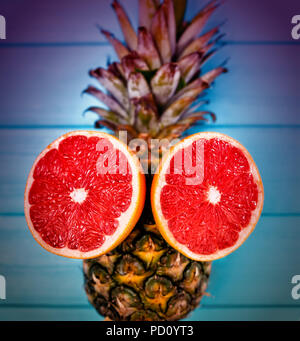 Colorful fresh halved ruby grapefruit arranged as eyes on a whole pineapple with vignette and toning in square format Stock Photo