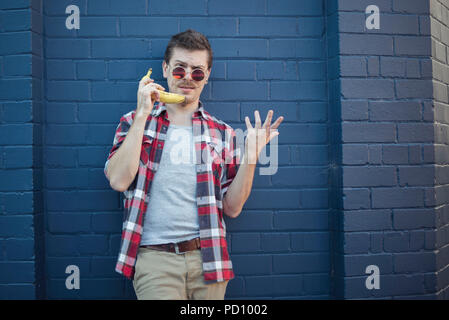 Young white man in hipster clothes, curly moustache, and vintage glasses, in front of a blue brick wall, using a banana as a phone. Stock Photo
