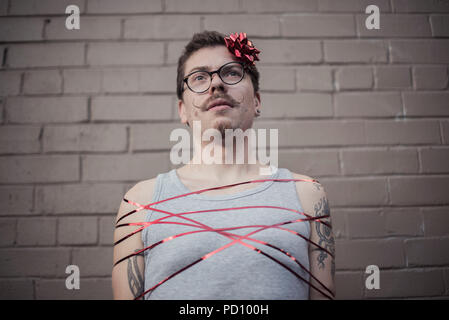 Young white man with tattoos wrapped in a red gift ribbon, with a red bow in the side of the head, curly moustache, and vintage glasses. Stock Photo