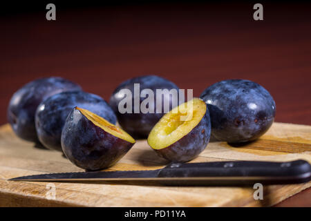 Fresh plums and one sliced plum with knife on a wooden board on dark red table. Stock Photo