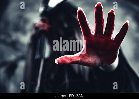 Witch showing bloody hand with reaper. Female demon angel in black clothes and hood on grunge wall background. Halloween day and Mystery concept. Fant Stock Photo