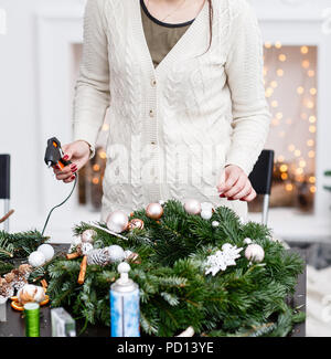 Woman decorated a Christmas wreath. Attaches toys and decor with glue gun. Hands close-up. Master class on making decorative ornaments. Christmas decor with their own hands. Flower shop Stock Photo