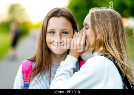 Two girl friends schoolgirl. In the summer in city. Tells a friend in the ear, a secret, really. The concept of surprise, best friends, gift. Emotion of happiness is the pleasure of a smile. Stock Photo
