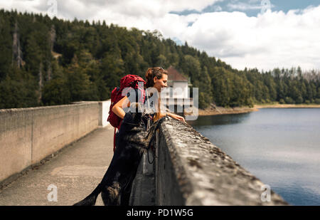 young woman with backpack and german shepherd dog puppy standing on a bridge in front of forest and lake Stock Photo