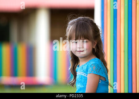 Portrait of little fashionable blond girl in blue dress, with gray eyes and beautiful long hair smiling in camera on bright blurred colorful backgroun Stock Photo