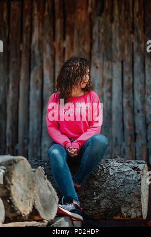 Graceful curly-haired woman having fun during photoshoot. Lovely tanned girl in pink shirt posing on wooden background Stock Photo