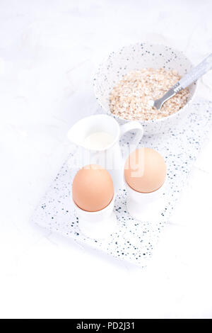 Boiled eggs for breakfast, oatmeal porridge and milk in white dishes on a light background. Delicious and healthy breakfast. Free space for text. Copy space Stock Photo