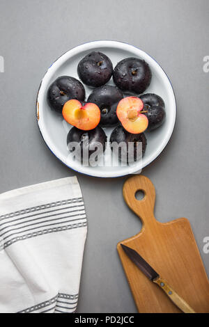 Dark red big plums whole and halved on white vintage enamel plate. Wooden cutting board knife white cotton towel on gray stone background. Minimalist  Stock Photo