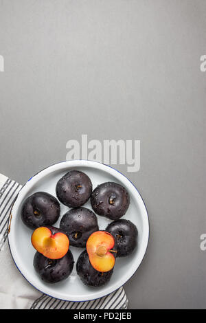 Dark red big plums whole and halved on white vintage enamel plate. White cotton towel on gray stone background. Minimalist style. Autumn fall harvest. Stock Photo