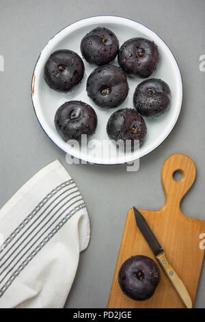 Dark red big plums on white vintage enamel plate. Wooden cutting board knife white cotton towel on gray stone background. Minimalist style. Autumn fal Stock Photo