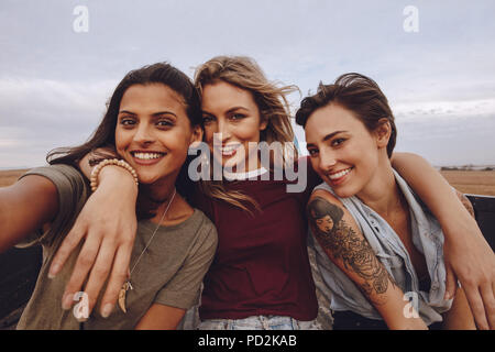 Beautiful women taking selfie while traveling by a pickup truck. Three young female friends posing for a selfie in a open back truck in country side. Stock Photo