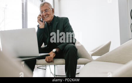 Mature businessman sitting in office lobby with a laptop using mobile phone. Senior male executive working in lobby. Stock Photo
