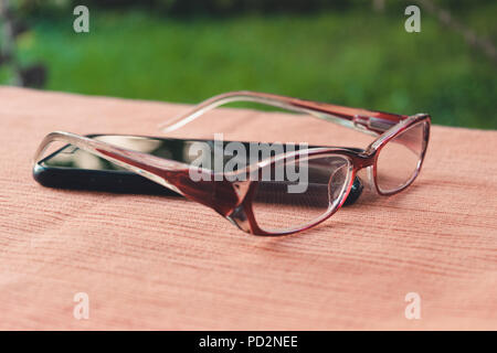 Close-up of eyeglasses and mobile phone on the table in the garden. Side view. Stock Photo